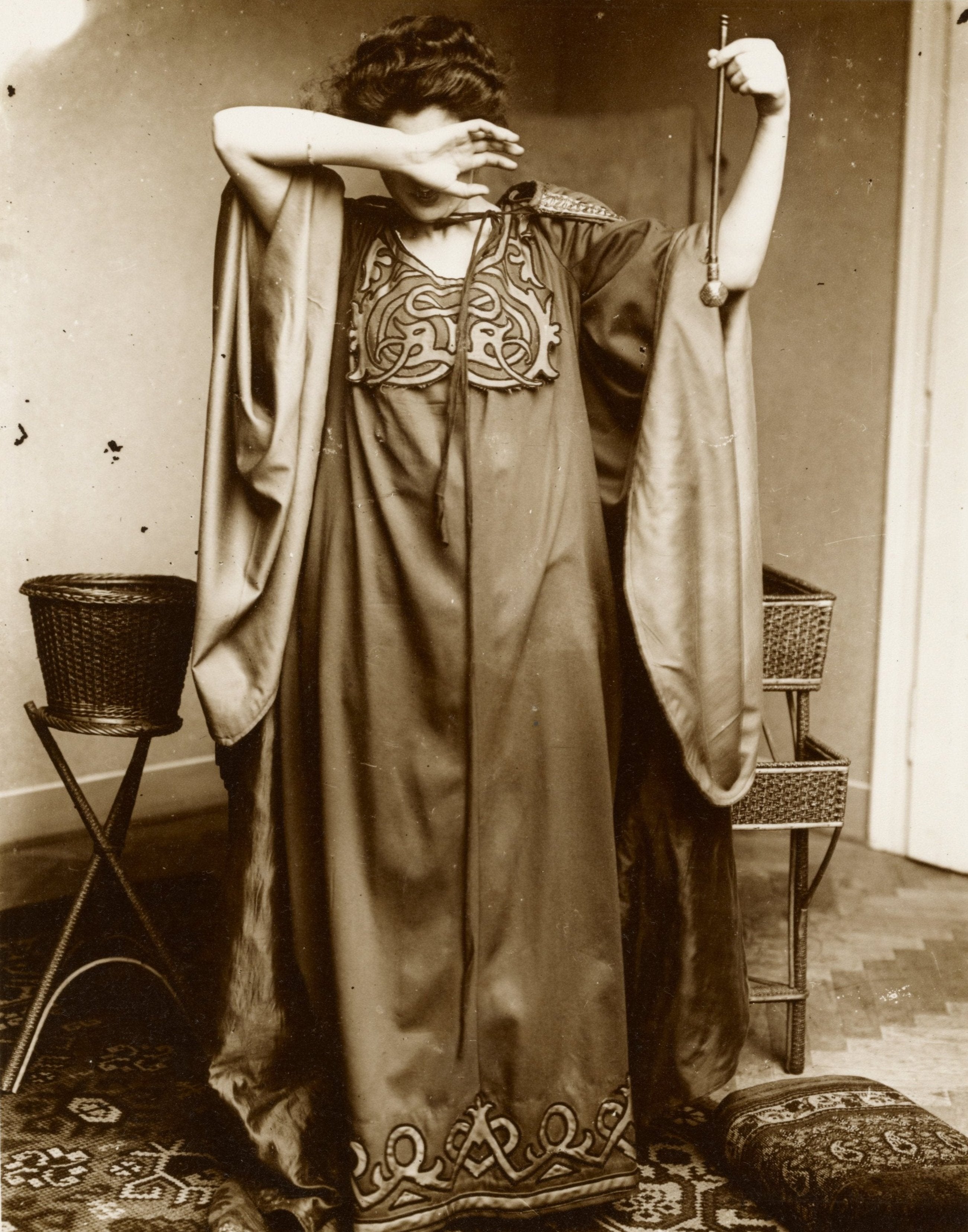 Black and white photo of a female model wearing a red dress with decorative patterns, posing with one hand over her face.