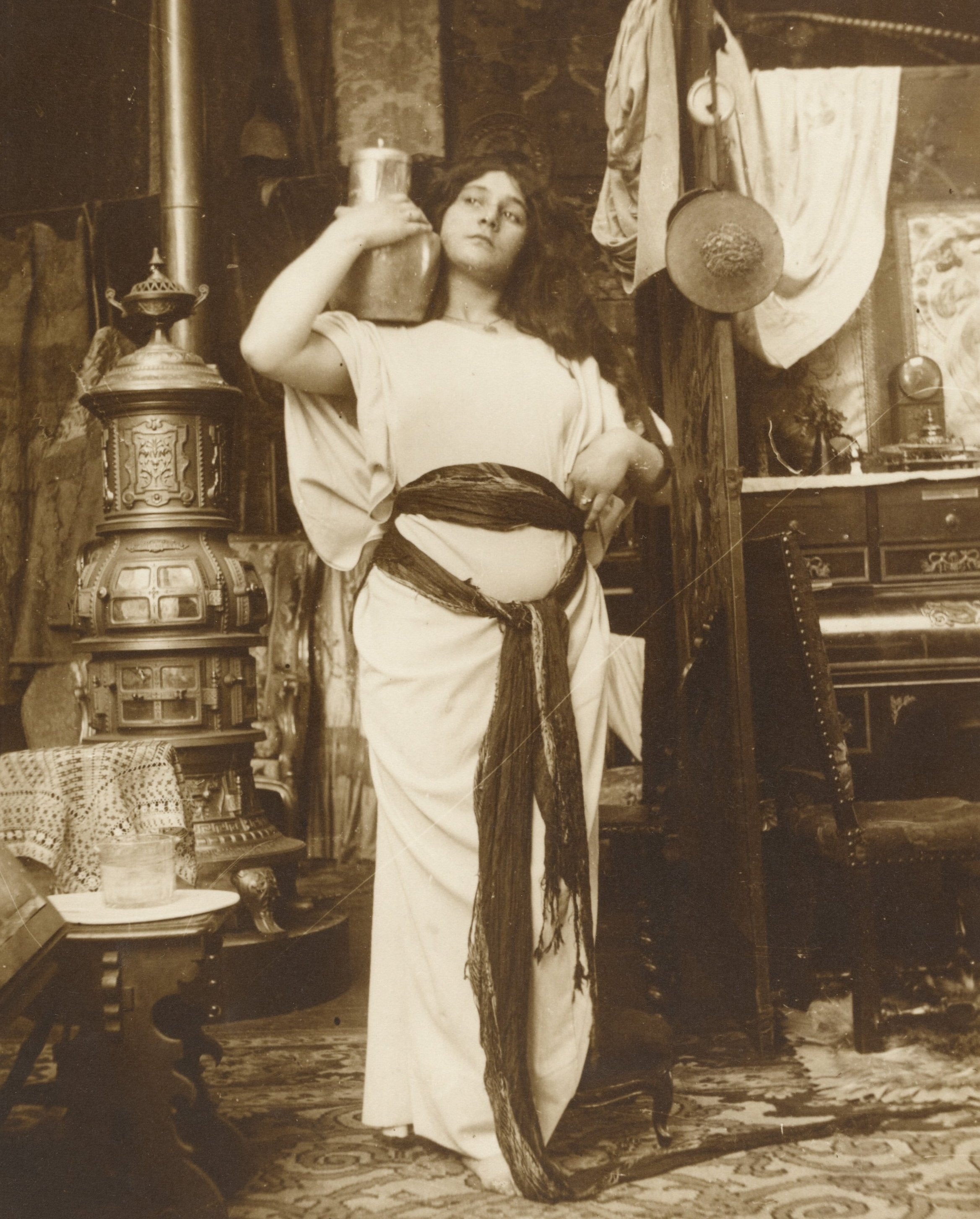 A black and white photo of a female model dressed in a white and red traditional outfit, standing in a room with a fireplace and decorative items.