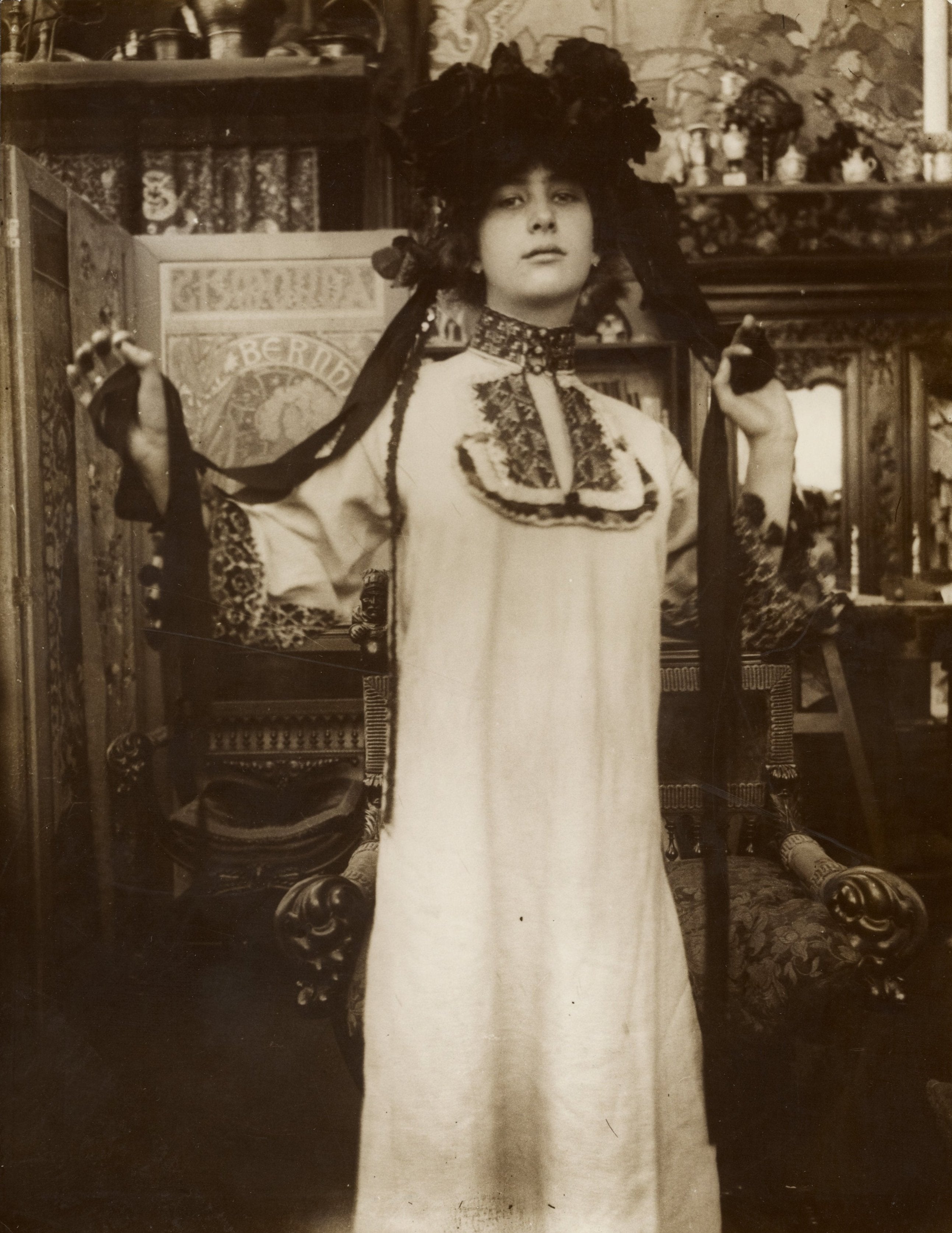 A black and white photograph of a female model wearing a long white robe with decorative elements, posed in a room with furniture and floral wallpaper.