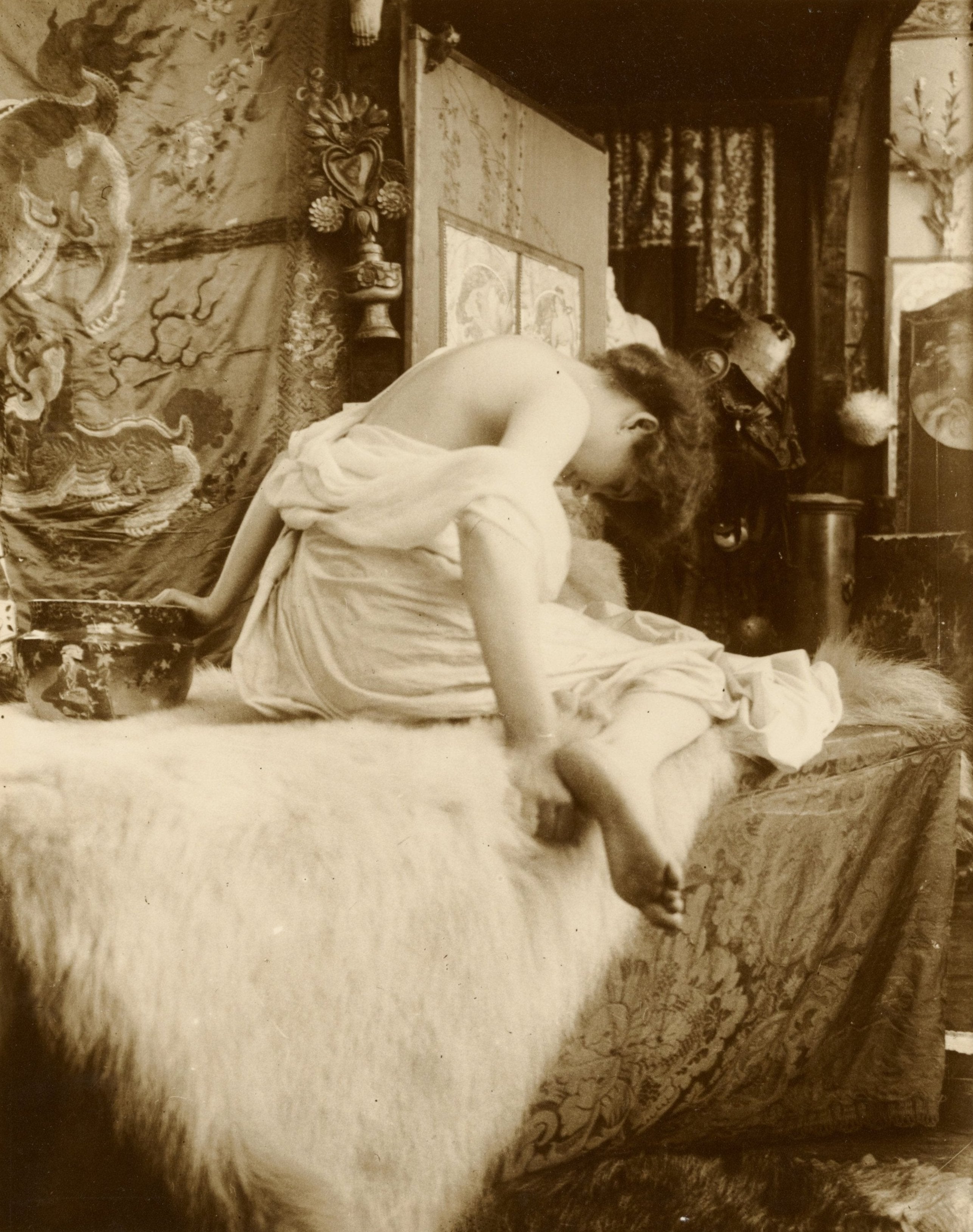 Black and white photograph of a female model seated on a bed, with a vintage decorative background.